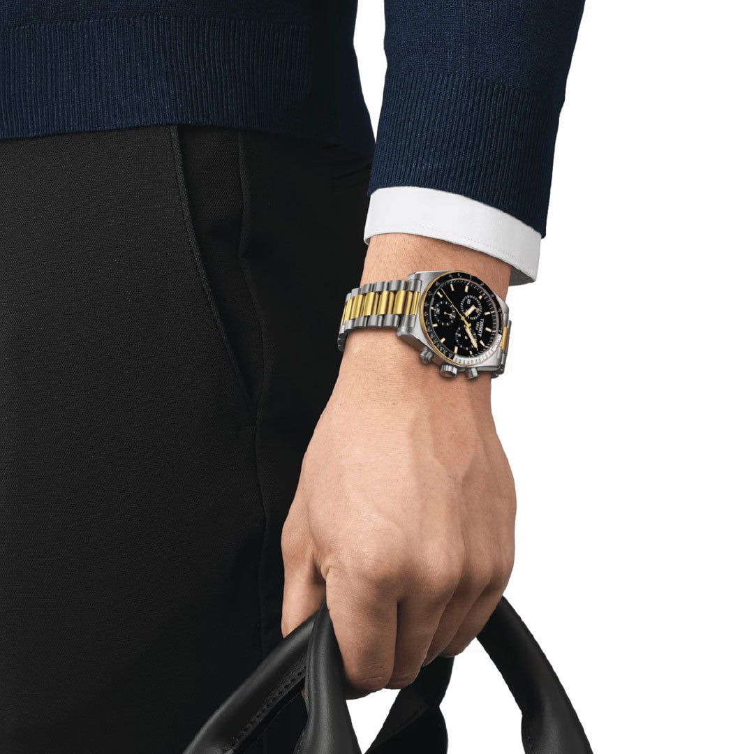 A person wearing a dark sweater and black pants holds a black leather bag. On their wrist, they sport the Tissot PR516 Chronograph Quartz 40mm Watch, showcasing a silver and gold design with a black dial. The sophisticated timepiece adds an elegant touch as they pose against the white background.