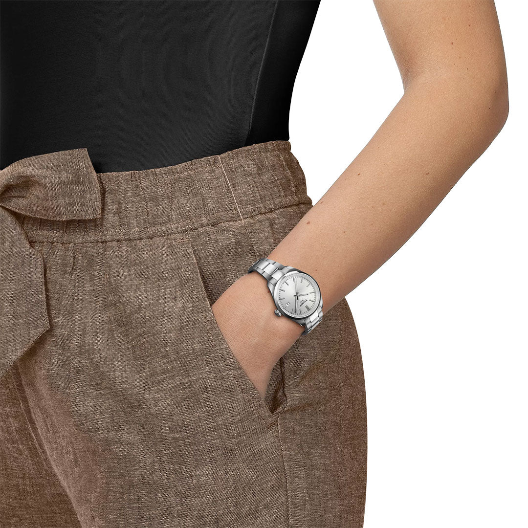 A person in brown high-waisted pants and a black top stands with one hand in their pocket, showcasing the Tissot PR100 Quartz 34mm Watch by Tissot with a white face on their left wrist.