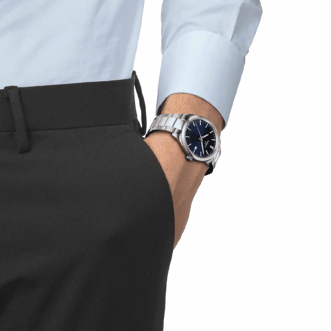 A person dressed in a light blue shirt and black pants, with their hand in the pocket, showcases a sleek Tissot PR100 Quartz 40mm Watch. The watch, crafted by Tissot, features a 316L stainless steel case and Swiss Quartz movement. Its black face is protected by a scratch-resistant sapphire crystal on their left wrist.