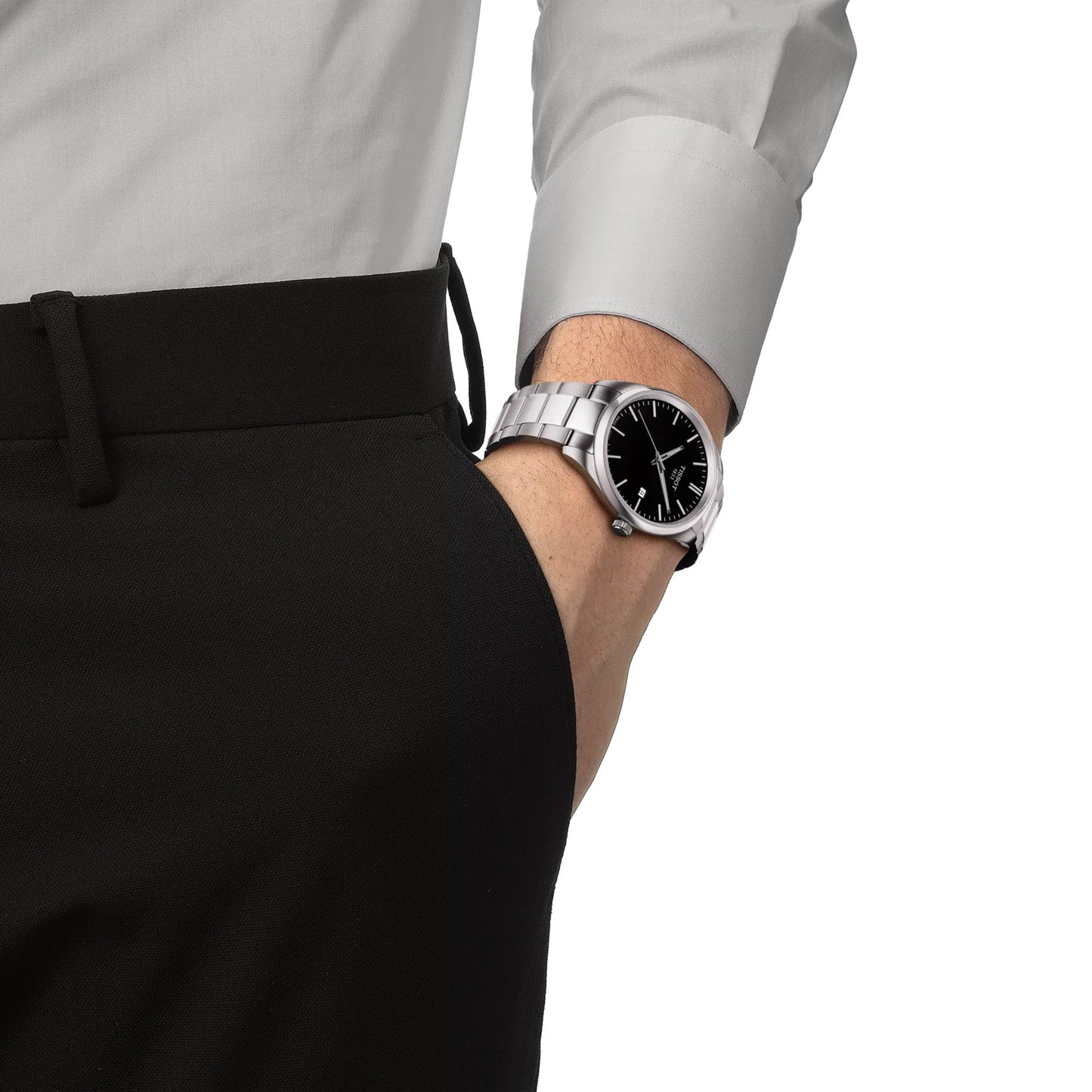 A person wearing a gray long-sleeve shirt and black pants has their hand in their pocket. On their wrist, they wear a Tissot PR100 Quartz 40mm Watch, featuring a black dial encased in stainless steel with scratch-resistant sapphire crystal.