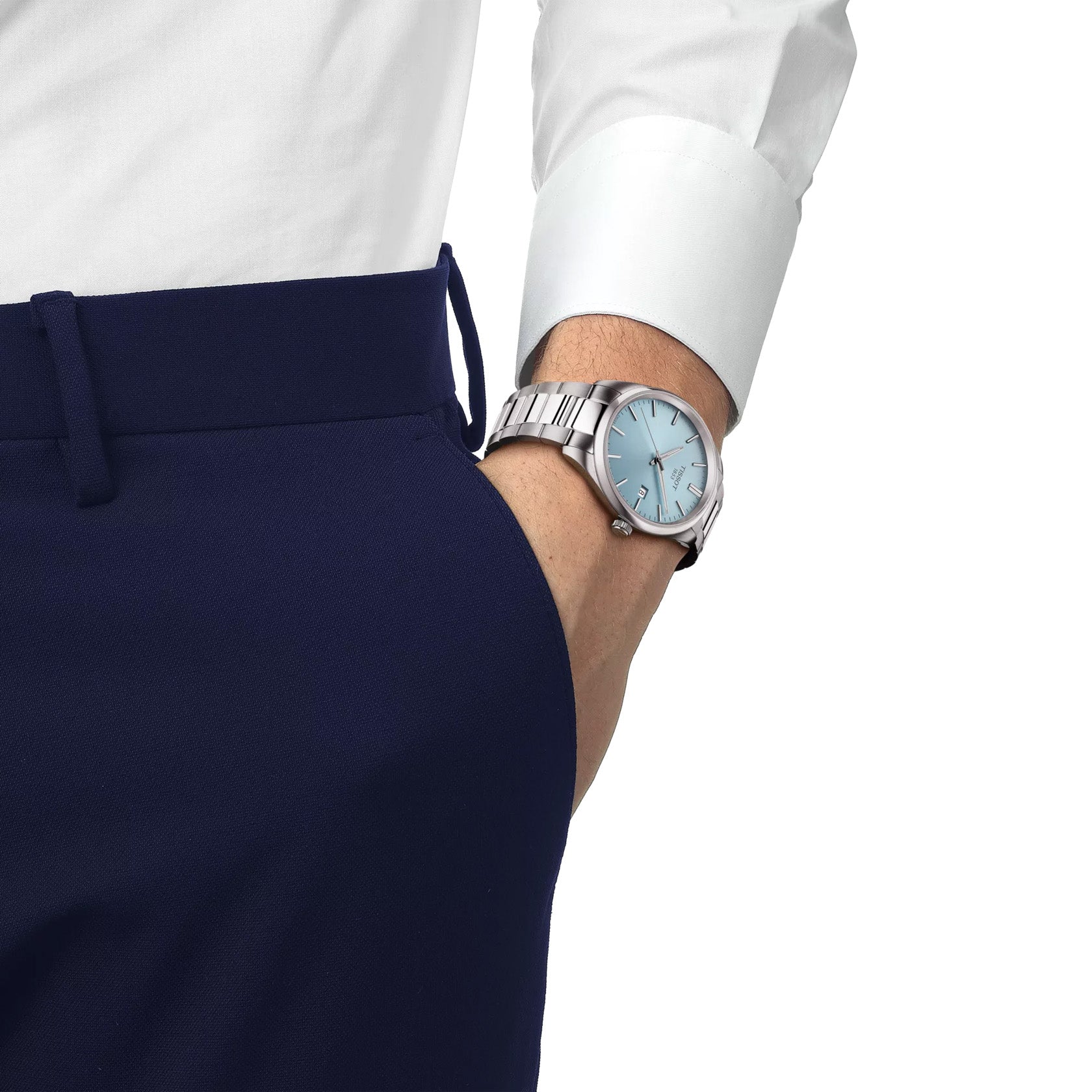 A person dressed in a white shirt and navy blue pants casually has their hand in their pocket while displaying a Tissot PR100 Quartz 40mm Watch, featuring a blue dial and stainless steel bracelet that highlights the sophistication of Swiss Made quartz movement.