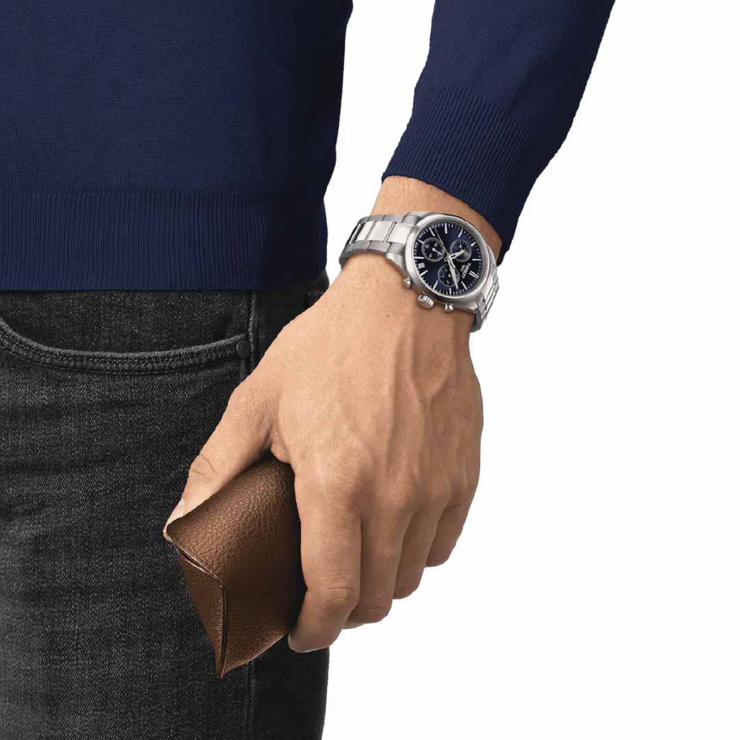 A person dressed in a blue sweater and gray jeans holds a brown leather wallet in one hand, while also wearing the Tissot PR100 Chronograph Quartz 40mm Watch on their wrist. This elegant silver timepiece features a black watch face with classic quartz chronographs for precision.