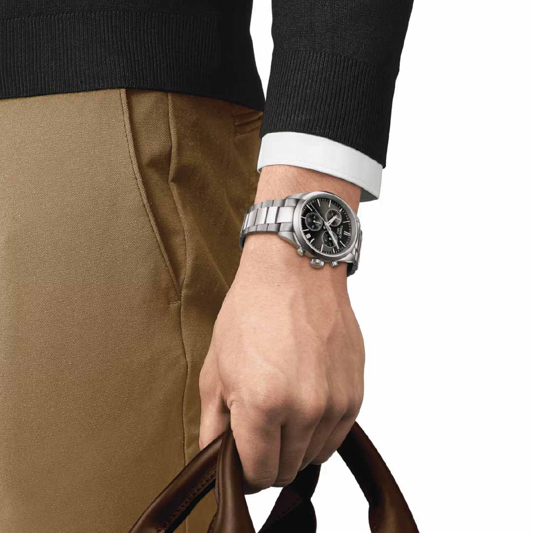 A person wearing a black sweater and brown pants holds a brown leather handle while their left wrist displays the Tissot PR100 Chronograph Quartz 40mm Watch, featuring a sporty design with a black dial. The image emphasizes the hand, watch, and clothing details, showcasing the sleek craftsmanship of quartz chronographs by Tissot with Super-LumiNova® coating.