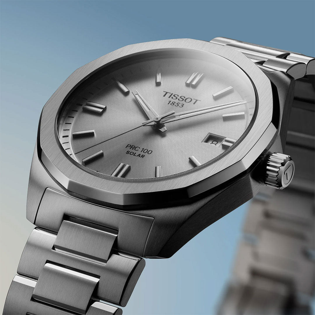 Close-up of the Tissot PRC 100 Solar Quartz 39mm Watch featuring a silver metal band, textured silver dial, date window, and index markers, set against a gradient blue background.