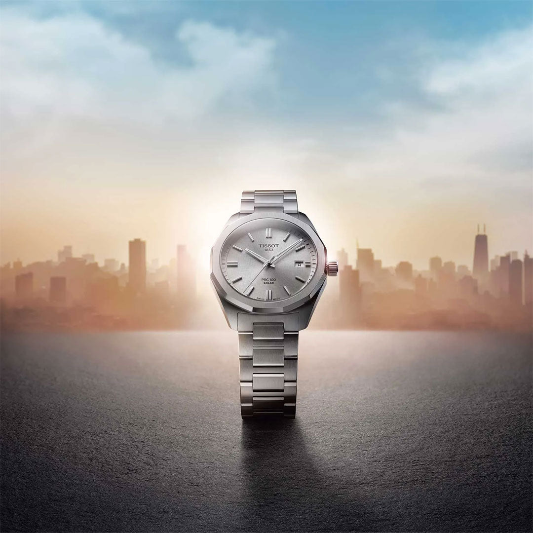 A Tissot PRC 100 Solar Quartz 39mm watch by Tissot, in silver, stands upright on a smooth surface with a blurred city skyline and a gradient sunrise sky in the background.