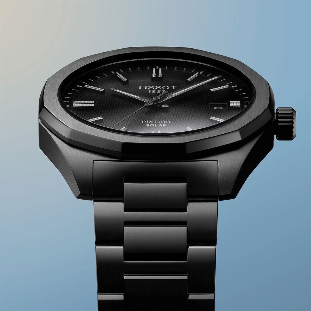 A Tissot PRC 100 Solar Quartz 39mm Watch with a black dial, metal bracelet, and minimalist markers is showcased against a gradient blue background.
