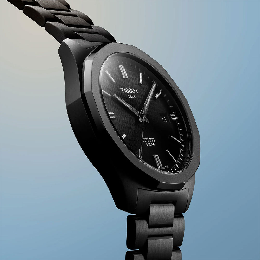A black Tissot PRC 100 Solar Quartz 39mm Watch with a metal band is angled against a blue and beige gradient background, showcasing the Tissot brand name, date feature, and solar markings that highlight its advanced technology.