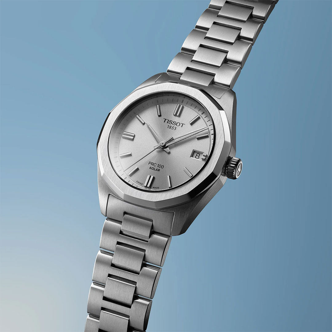 The Tissot PRC 100 Solar Quartz 34mm Watch, featuring a silver dial, metal band, date window, and minimalist hour markers, is shown against a blue gradient background.