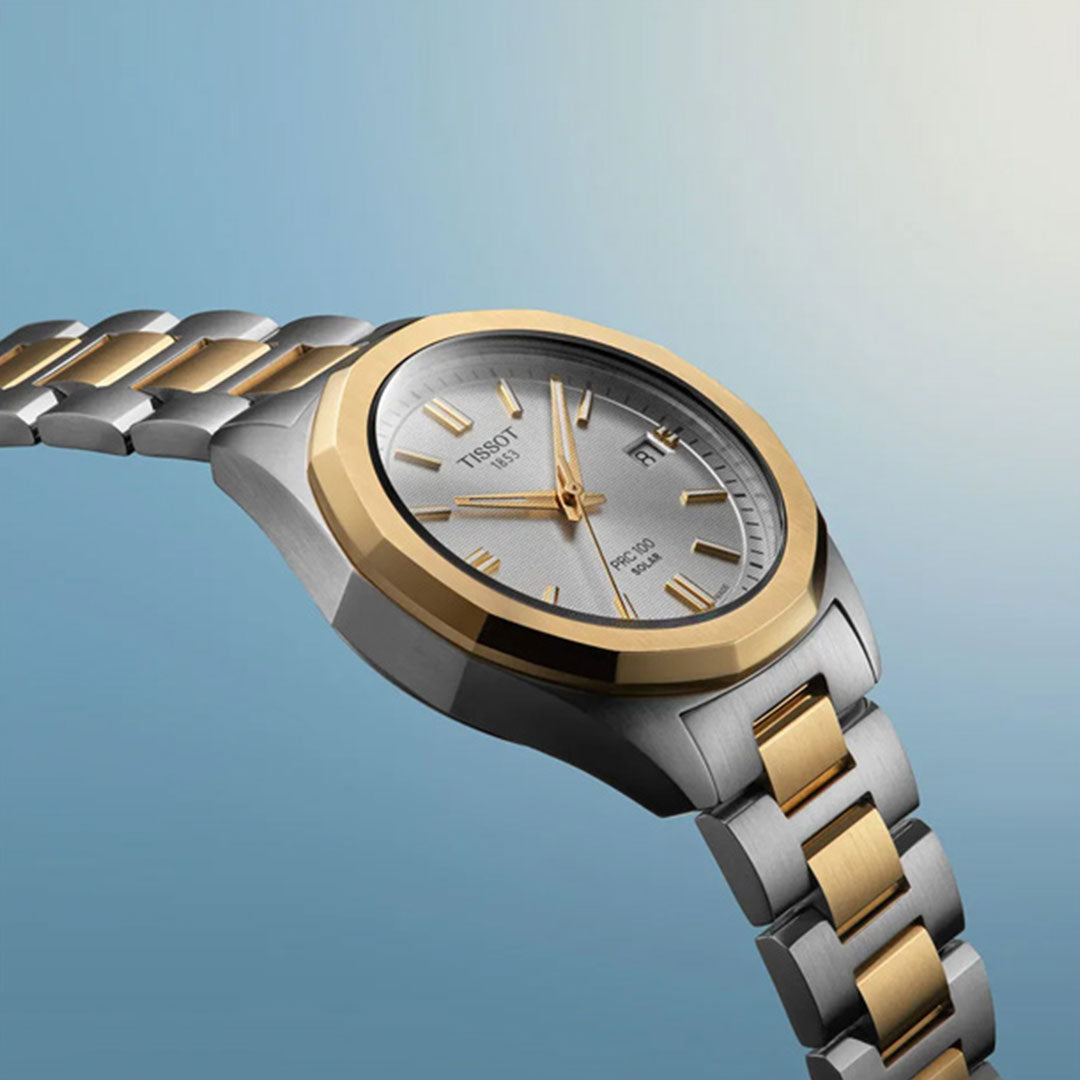 A close-up of the Tissot PRC 100 Solar Quartz 34mm Watch, featuring a silver and gold metal band, round silver face, gold hour markers, and date display by Tissot, set against a gradient blue background.