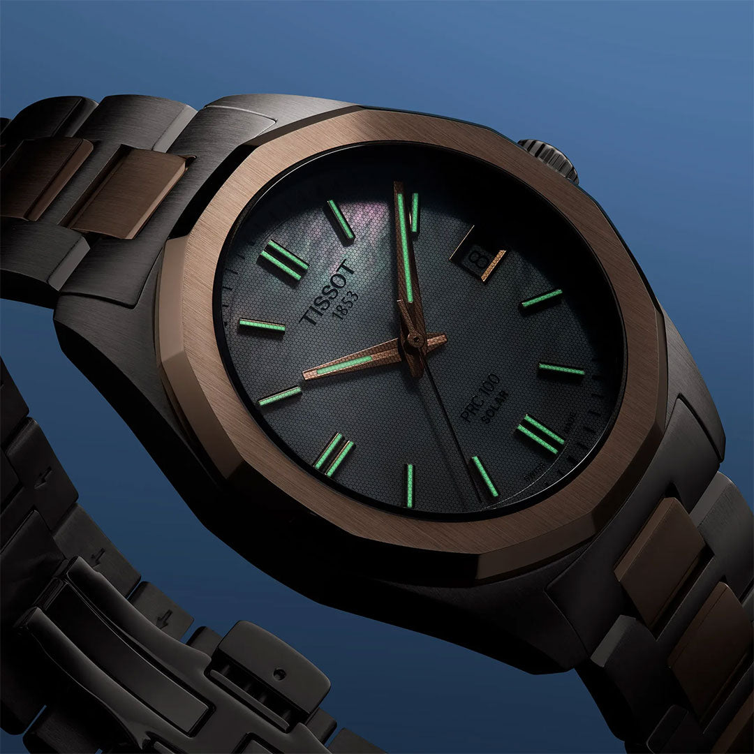 A close-up of the Tissot PRC 100 Solar Quartz 34mm Watch showcases its dark metallic band, bronze bezel, and textured black dial with luminous green markers and hands, powered by solar quartz, set against a blue gradient background.