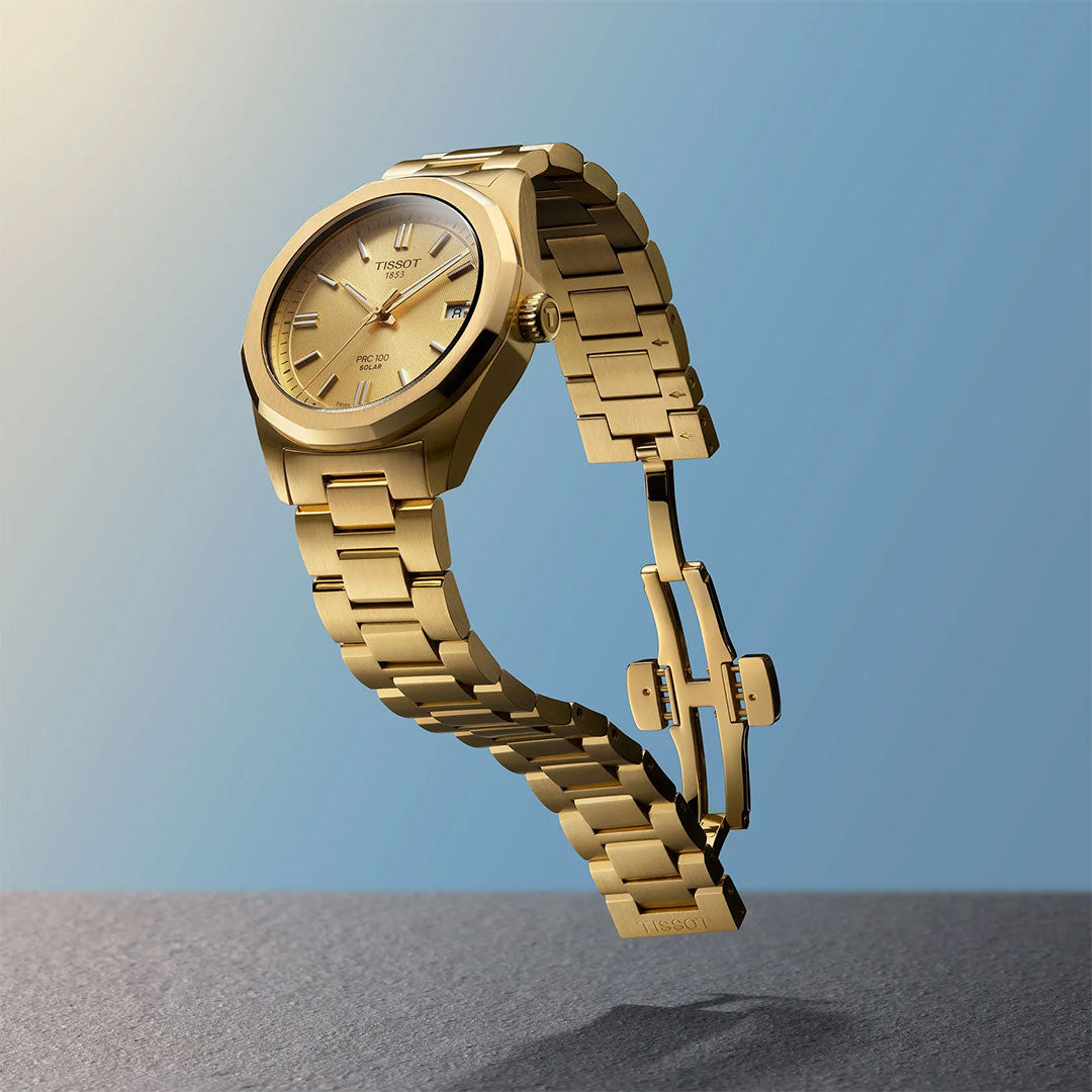 A Tissot PRC 100 Solar Quartz 34mm Watch by Tissot, featuring a gold metal band and open clasp, is showcased suspended against a blue gradient background above a textured gray surface.