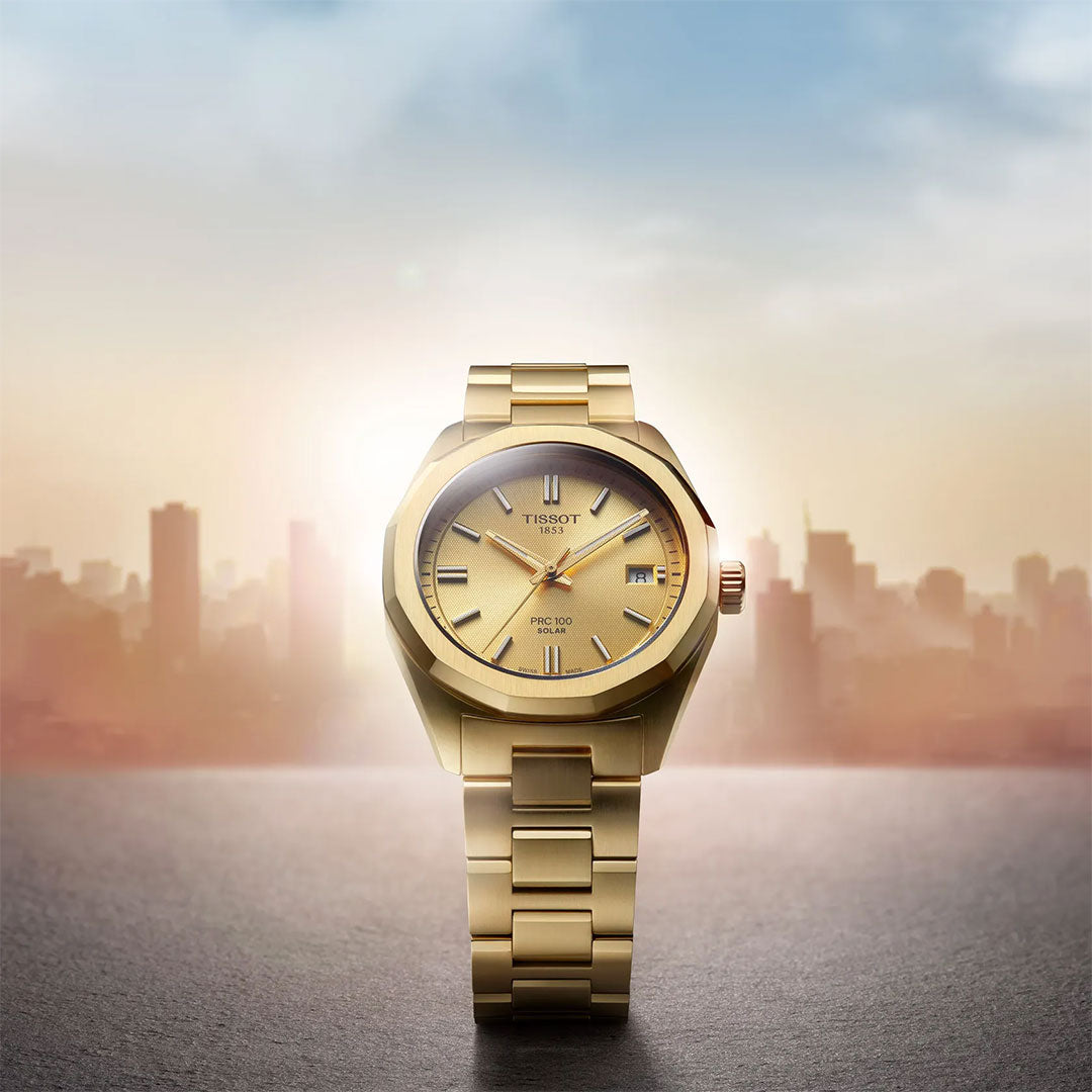 A Tissot PRC 100 Solar Quartz 34mm Watch is shown upright on a surface, its shiny gold-toned finish and elegant design highlighted against a blurred city skyline and bright sun in the background.