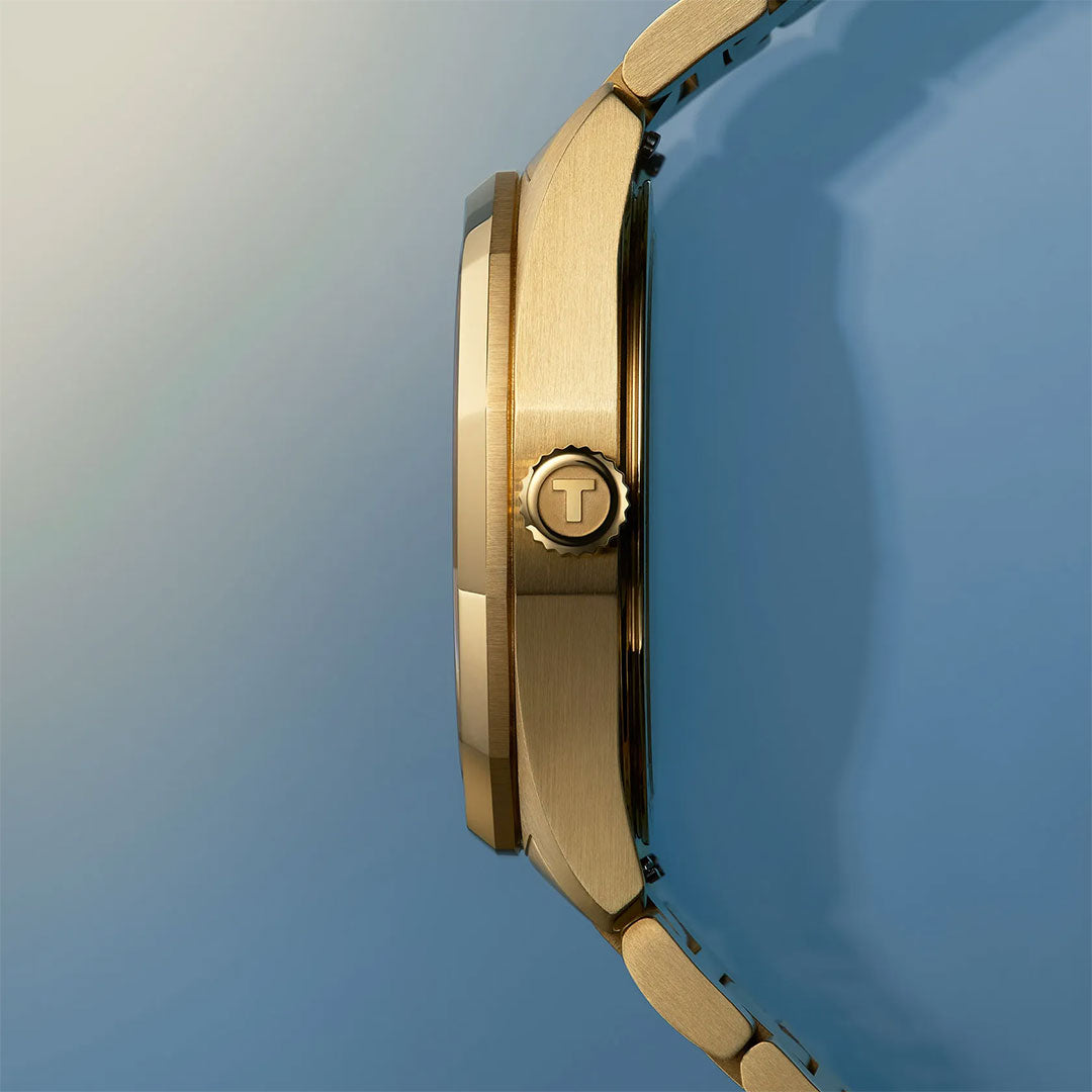 Side view of the Tissot PRC 100 Solar Quartz 34mm Watch in gold against a gradient beige to blue background, highlighting its round case and signature "T" crown—an elegant solar-powered quartz timepiece by Tissot.