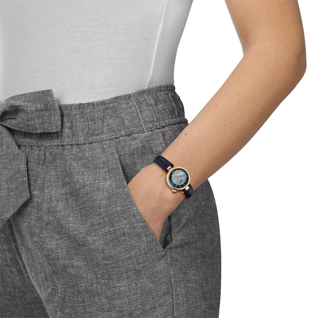 A person in gray high-waisted pants with a bow and a white top has their hand in their pocket, revealing a Tissot Desir Automatic 28mm Watch with a blue mother-of-pearl dial and gold accents on their left wrist.