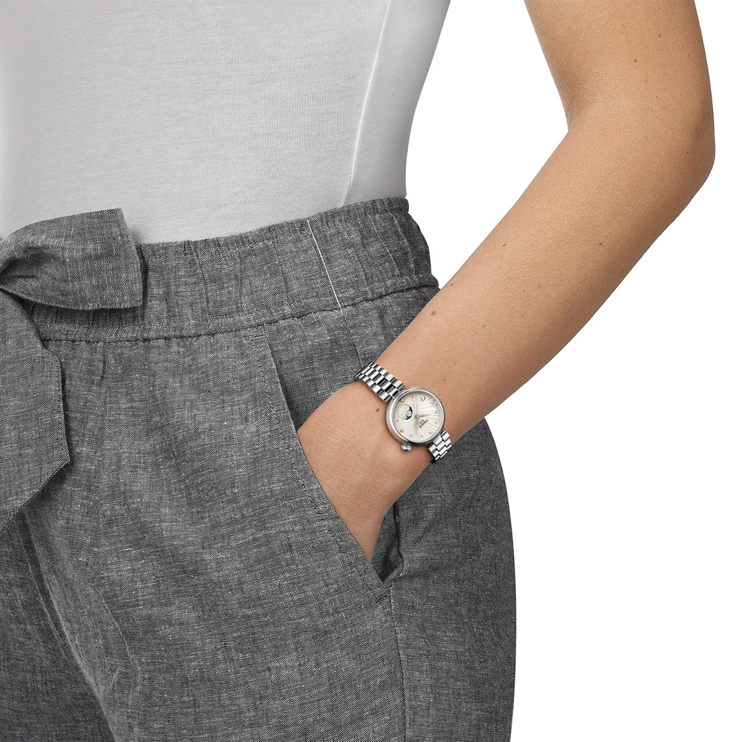 A person dressed in gray high-waisted pants and a white top has their right hand in their pocket, showcasing the Tissot Desir Moonphase Quartz 29.6mm Watch by Tissot on their wrist.