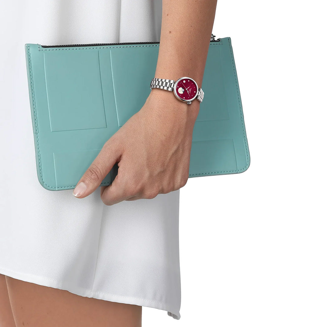 A person in a white outfit holds a light blue clutch and wears a silver Tissot Desir Moonphase Quartz 29.6mm Watch by Tissot with a striking red dial on their left wrist.