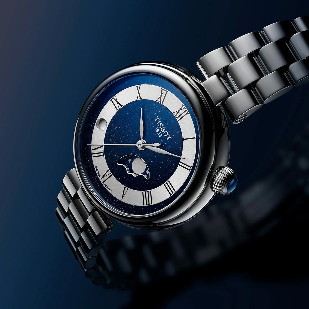 A Tissot Desir Moonphase Quartz 29.6mm watch by Tissot, featuring a silver and blue dial with Roman numerals, a moonphase complication, and a stainless-steel bracelet, displayed against a dark blue gradient background.