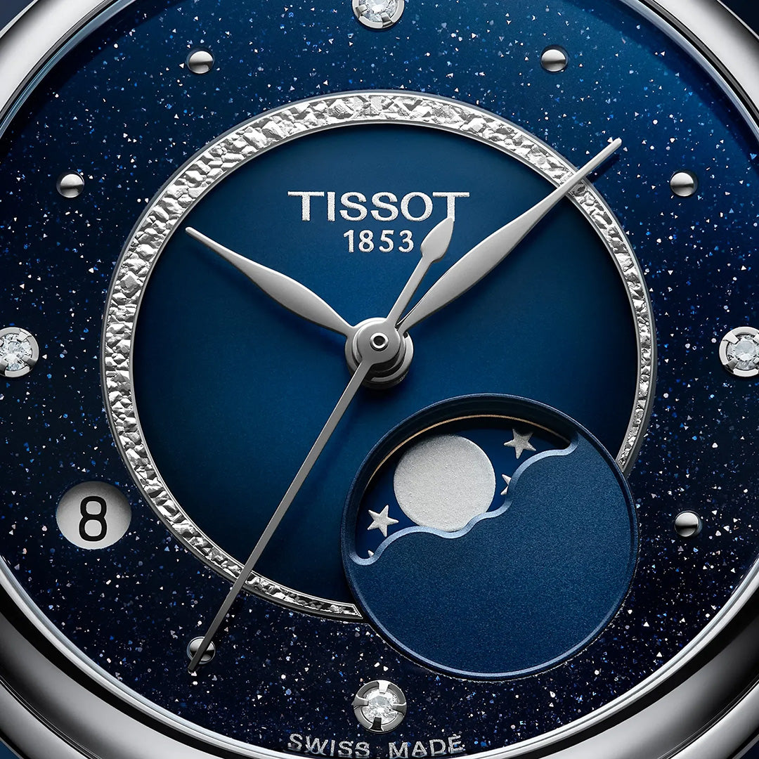 Close-up of the Tissot Desir Moonphase Quartz 29.6mm Watch face set against a blue, starry background, featuring silver hands, diamond hour markers, and a moon phase display with moon and stars. The date display shows the number 8. This elegant Tissot timepiece is complemented by its diamond stainless-steel bracelet, adding sophistication to this stunning moonphase watch.