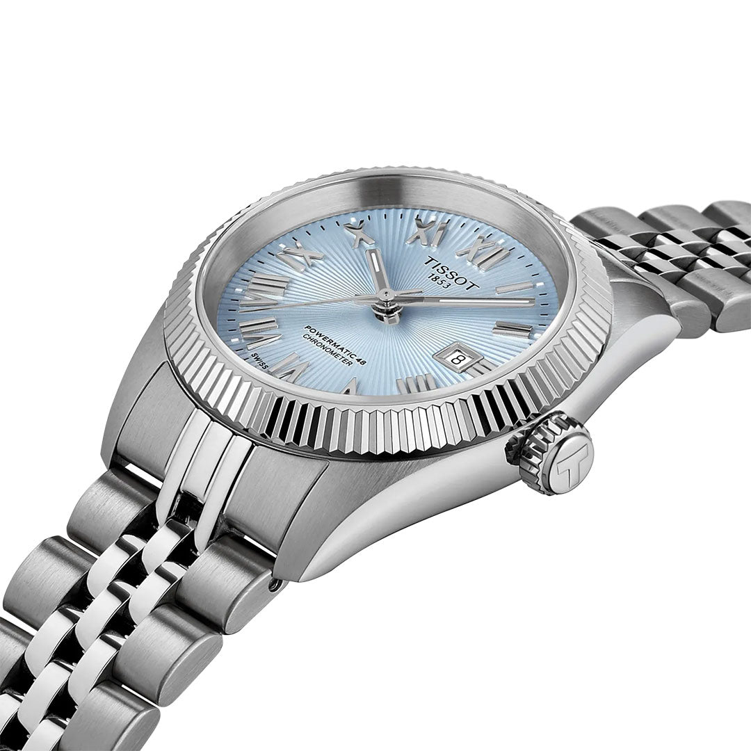 A silver Tissot Ballade Powermatic 80 Automatic 30mm Watch features a light blue textured dial, Roman numerals, date at 3 o’clock, fluted bezel, metal link bracelet, and COSC-certified movement.