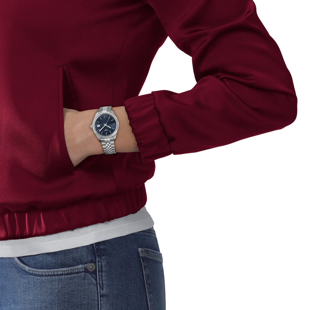 A person in a maroon jacket and blue jeans has their left hand in their pocket, showcasing a Tissot Ballade Quartz 34mm Watch with a silver case, guilloché dial, and blue face from Tissot.