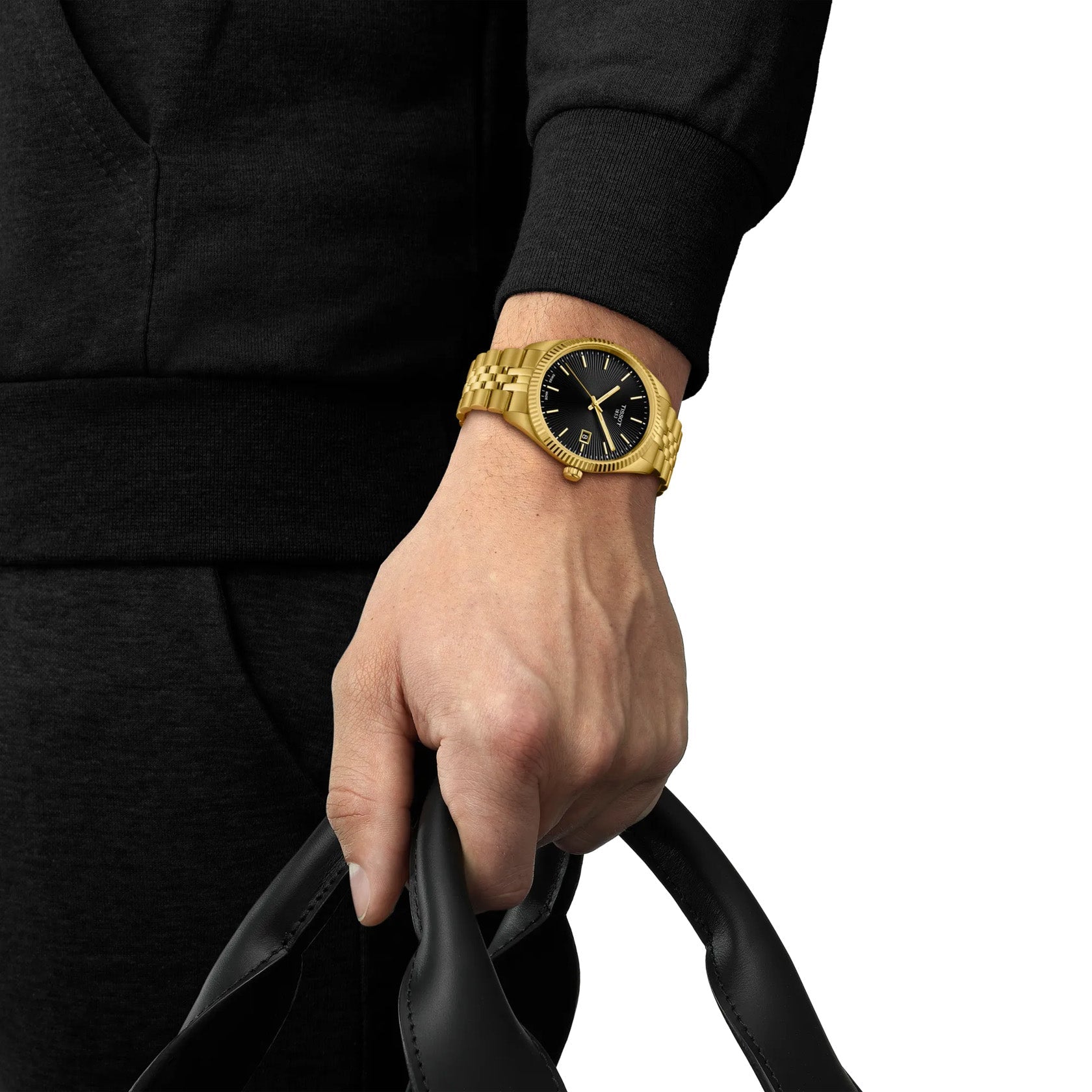 A person in black holds a black leather bag and wears the Tissot Ballade Quartz 40mm Watch. The gold watch with guilloché dial and markers adds an intricate touch against their dark outfit.