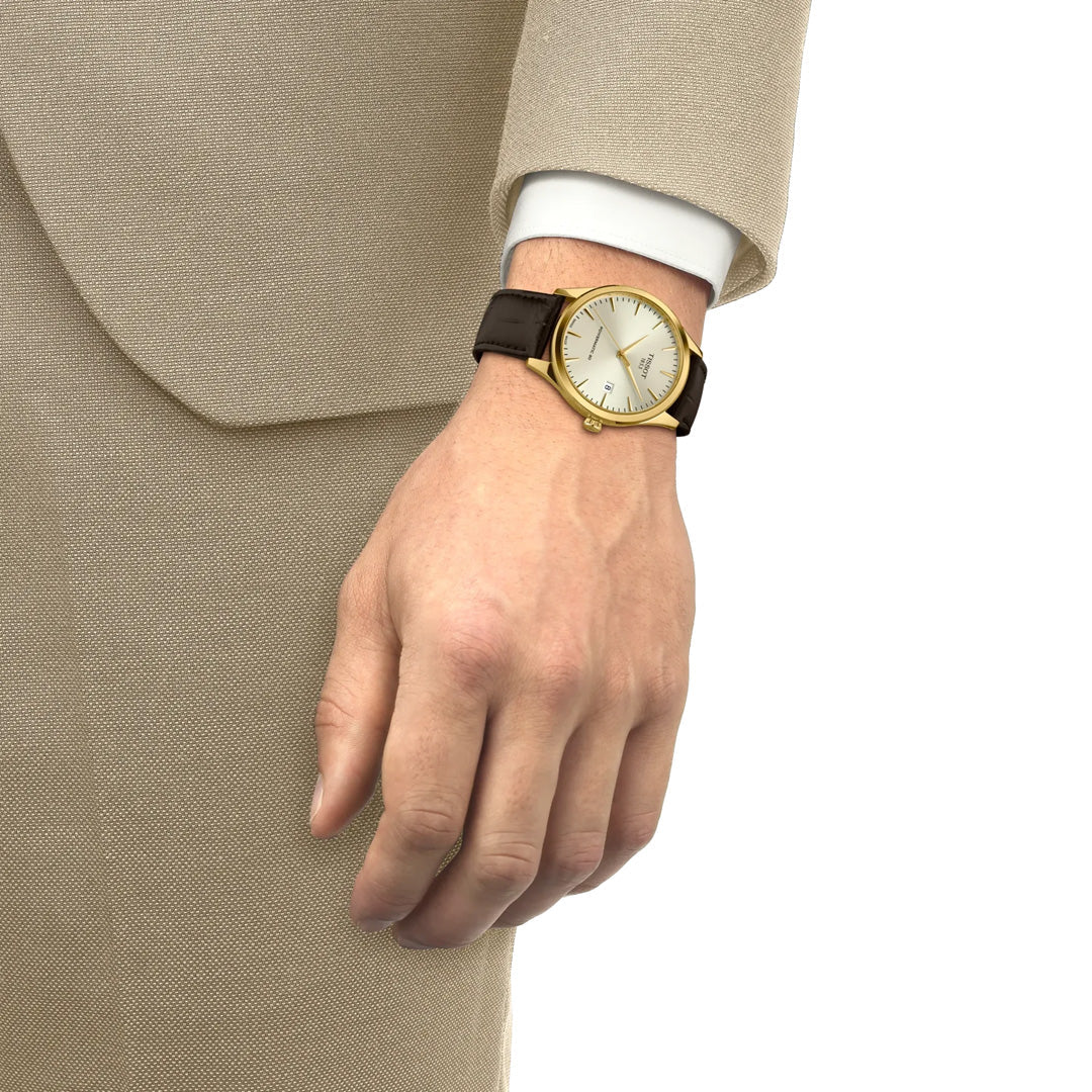 A person in a beige suit is shown from the shoulder down, displaying a Tissot Classic Dream Swissmatic 40mm watch with a brown leather strap and scratch-resistant sapphire crystal on their left wrist.