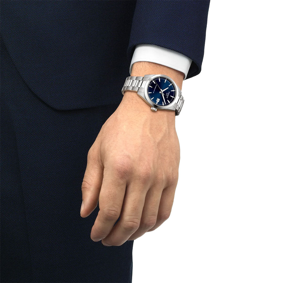 A person in a dark blue suit and white shirt is shown from the wrist down, wearing a Tissot Gentleman Powermatic 80 Automatic 38mm Watch with a blue dial and stainless steel bracelet on their left wrist.