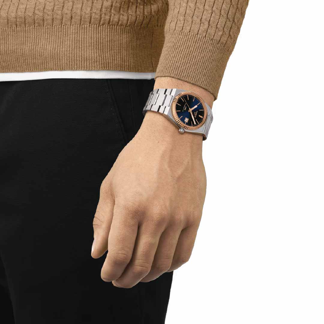 A person dressed in a brown sweater and black pants is displaying the Tissot PRX 18K Gold Powermatic 80 Automatic 40mm Watch, which features a sleek metallic band and a striking black and gold face. The watch is highlighted by an exquisite 18K rose gold bezel and powered by a Swiss automatic movement, with their arm resting casually at their side.