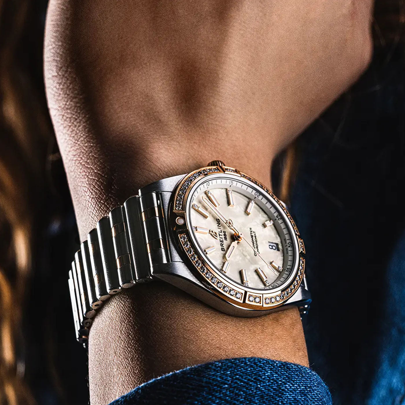 A close-up of a wrist showcasing the luxury BREITLING Chronomat Automatic 36, featuring a sophisticated silver and gold metal band. The watch face is elegantly white with gold hour markers and hands, complemented by a bezel decorated with small sparkling stones. The background appears slightly blurred.