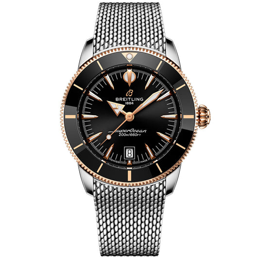 The Breitling Superocean Heritage B31 Automatic 42 features a black dial, gold accents, date at 6 o'clock, silver and gold hands and markers, a black bezel with a gold edge, and a stainless steel mesh bracelet.