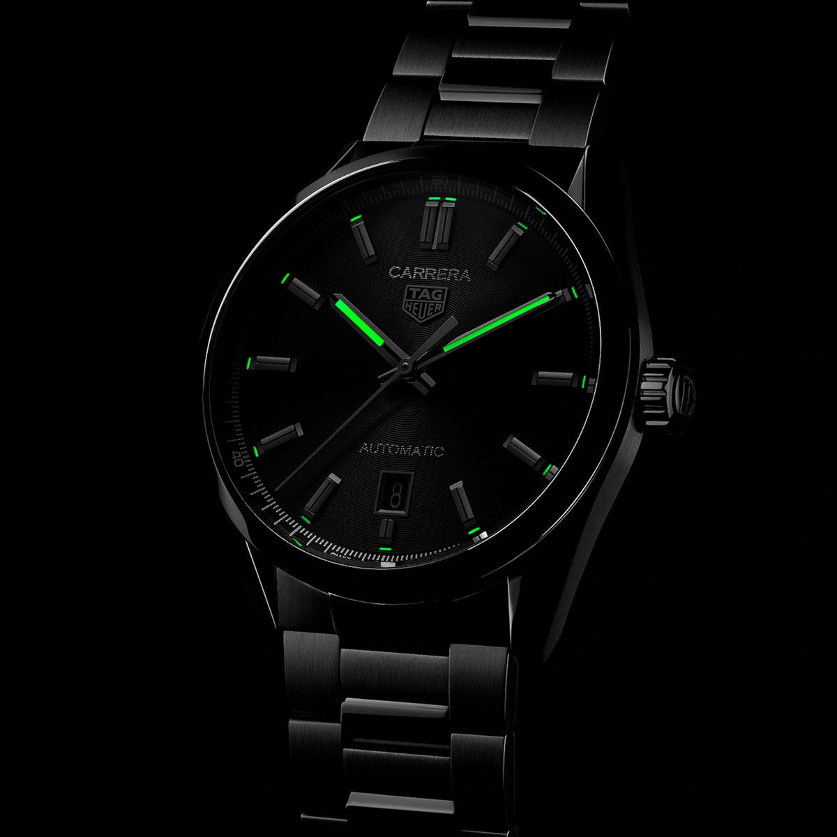 The TAG Heuer Carrera Date Automatic 39mm watch by TAG Heuer is displayed with a black dial, green luminescent hour markers and hands, and a matching metal bracelet, set against a dark background.