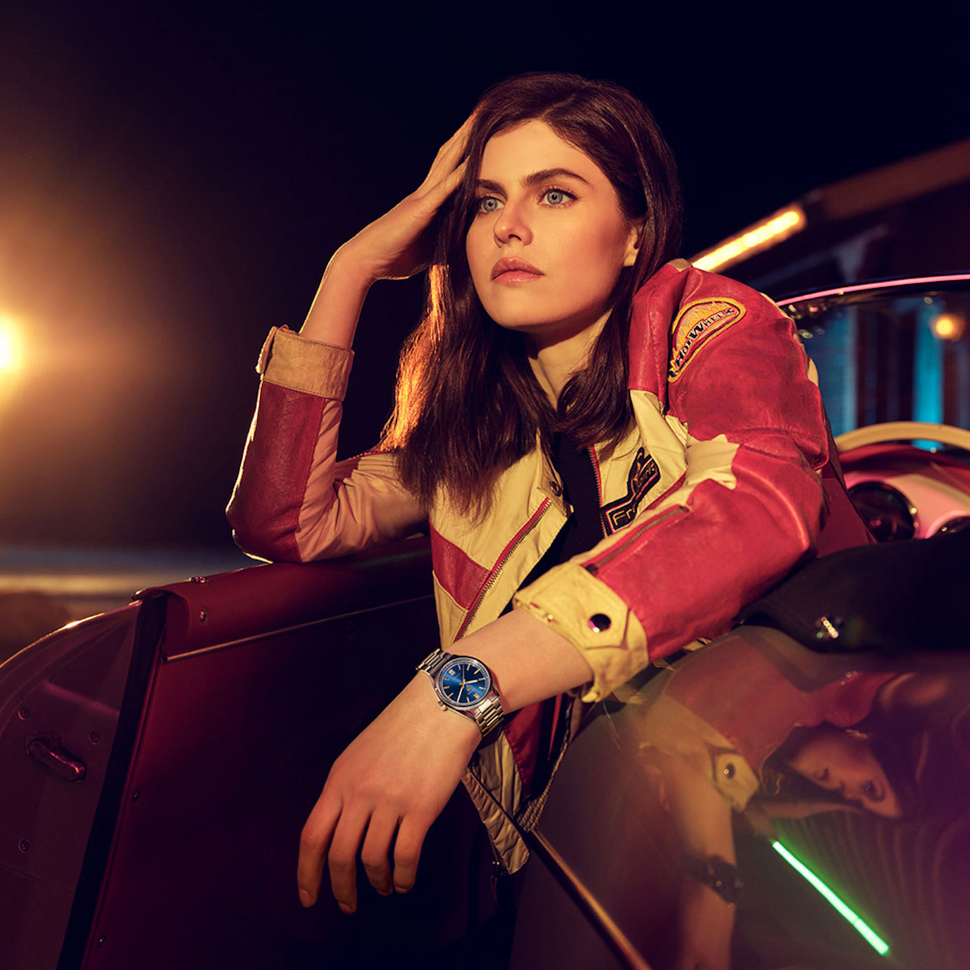 A woman in a red and beige jacket leans on a car door at night, gazing into the distance with her head resting on her hand. She wears the TAG Heuer Carrera Date Automatic 36mm watch by TAG Heuer, its face illuminated by dramatic, warm lighting that accentuates the refined elegance of its automatic movement.