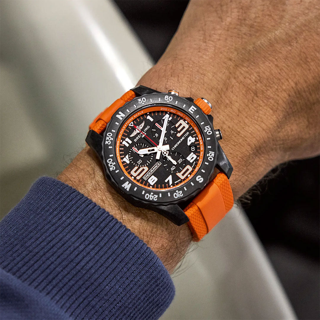 A person with medium skin tone, dressed in a blue long-sleeve shirt, wears a Breitling Professional Endurance Pro 44 featuring a black dial with white numerals and an orange rubber strap.