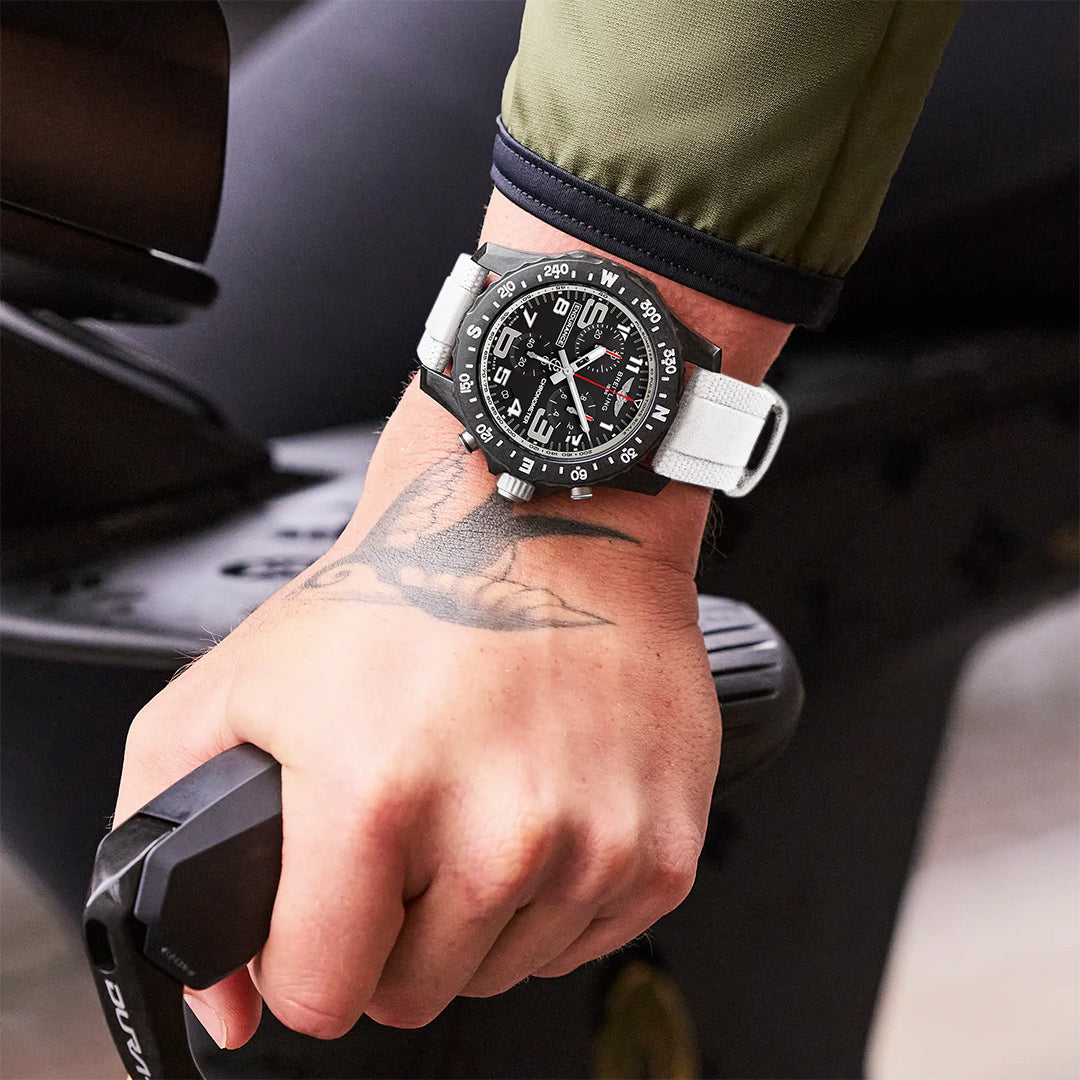 A person with a bird tattoo on their wrist wears a Breitling Endurance Pro 44 athleisure watch while gripping the handlebar of a black scooter or motorcycle.