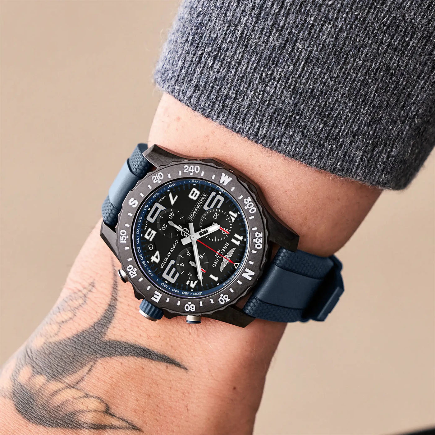 A person wears a BREITLING Endurance Pro 44 watch, featuring a blue strap and black dial with white numerals and compass markings. This timepiece is crafted from durable Breitlight® material. Their wrist, which displays a tattoo, emerges from under a gray, textured sleeve.