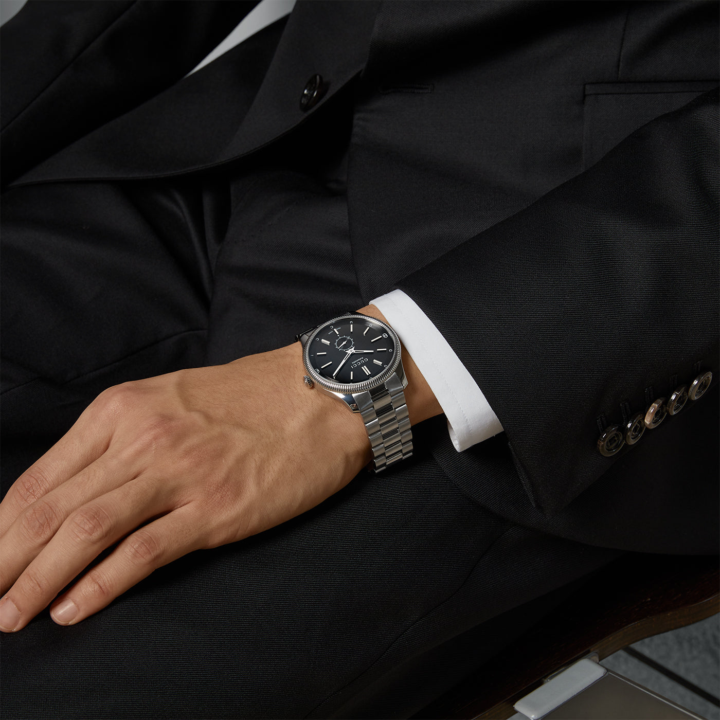 A person in a black suit sits with their hand resting on their lap, showcasing the Gucci G-Timeless Automatic 40mm Watch from GUCCI, featuring a black dial. The stainless steel bracelet elegantly complements the suit, which is adorned with detailed buttons on the sleeve.