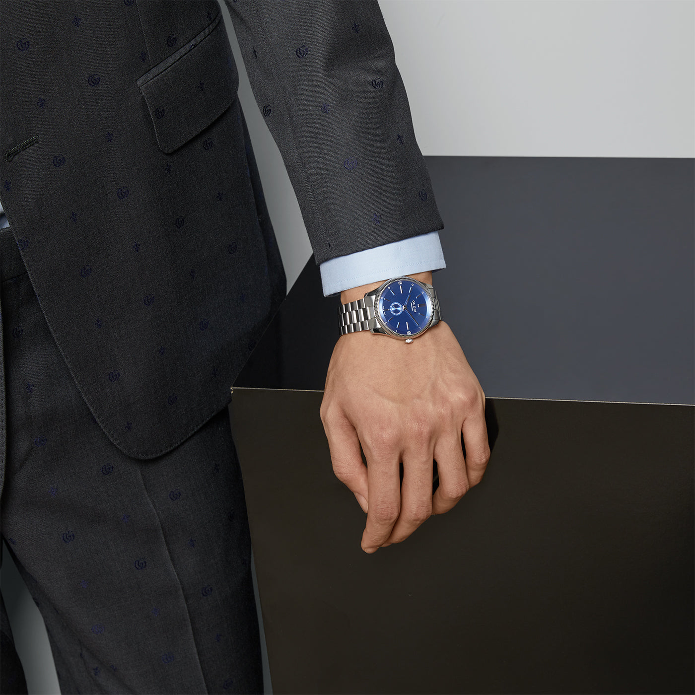 A person clad in a dark suit featuring subtle patterns rests their hand on a black surface. The elegant touch is provided by the sleek GUCCI Watch, specifically the Gucci G-Timeless Automatic 40mm Watch, with its captivating blue face and steel bracelet adorning their wrist.