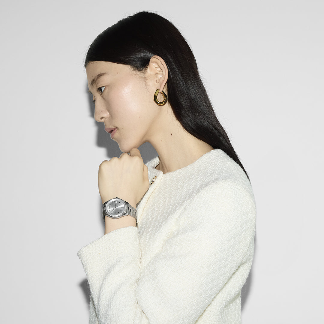 Profile view of a person with long, dark hair, wearing a textured white top, hoop earrings, and a Gucci G-Timeless Quartz 38mm Watch by GUCCI Watch, posed against a plain light background.