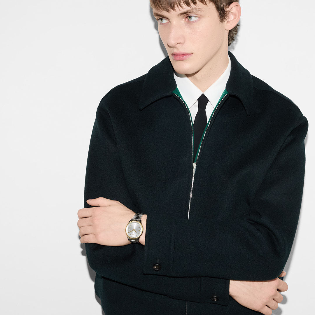 A young man with light skin and short brown hair stands against a white background, wearing a black zip-up jacket over a white shirt and black tie. He crosses his arms, wearing a GUCCI G-Timeless Quartz 38mm Watch, and looks off to the side.