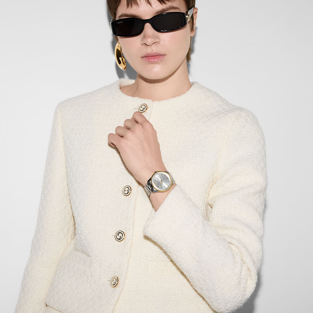 A person wearing a textured white jacket, black sunglasses, large gold earring, and a GUCCI G-Timeless Quartz 38mm Watch poses against a plain light background.
