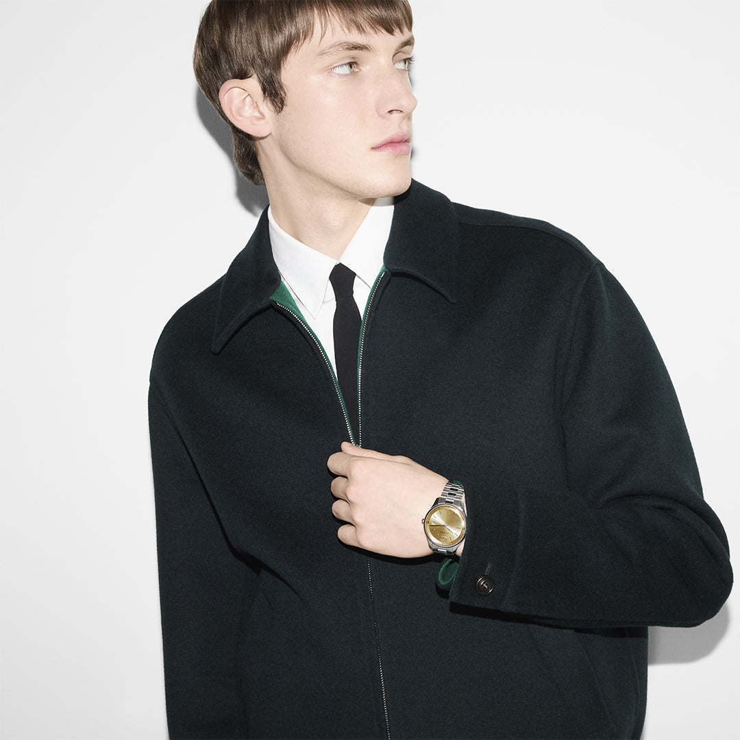 A person wearing a dark coat over a white shirt and black tie looks to the side, clutching the front of their coat with one hand. Their Gucci G-Timeless Quartz 38mm Watch, featuring an elegant gold-plated dial, stands out against the plain white background.