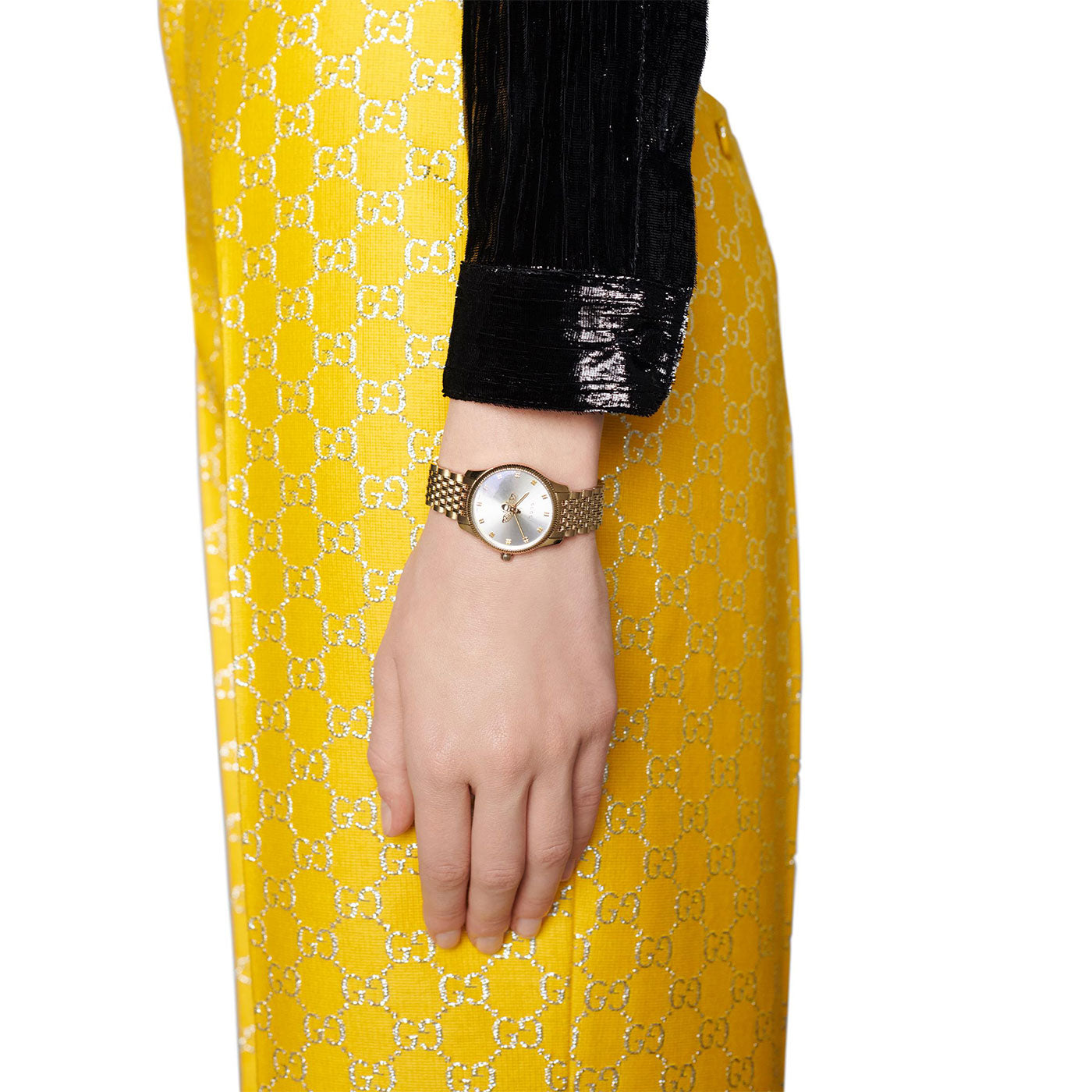 A person dressed in a shiny black top and yellow logo-patterned pants is showcasing the Gucci G-Timeless Quartz 29mm Watch. The round-faced watch, featuring a textured band, perfectly complements the luxurious style of their outfit.