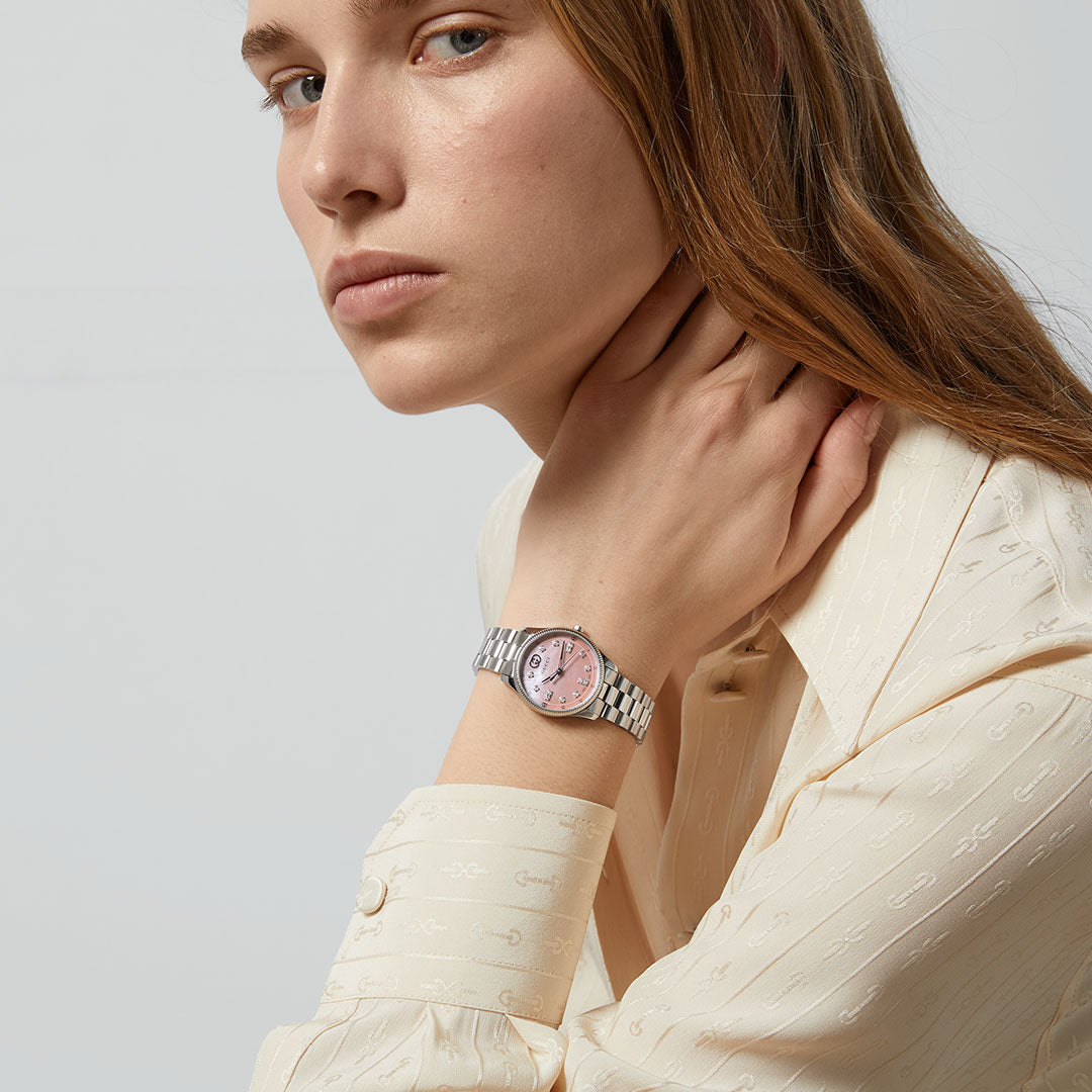 A woman with long light brown hair, in a cream shirt, rests her hand on her neck and looks at the camera. On her left wrist, she wears a GUCCI Watch: Gucci G-Timeless Quartz 29mm Watch with a pink face.