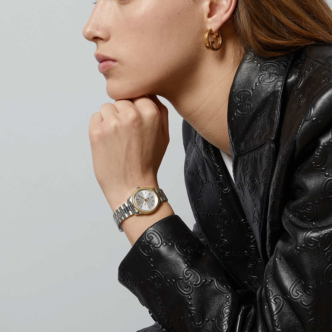 A person wearing a black patterned leather jacket and gold hoop earrings rests their chin on their hand, showcasing a GUCCI Watch from the G-Timeless Quartz 29mm collection. The elegant mix of silver and gold beautifully complements the plain light gray background.