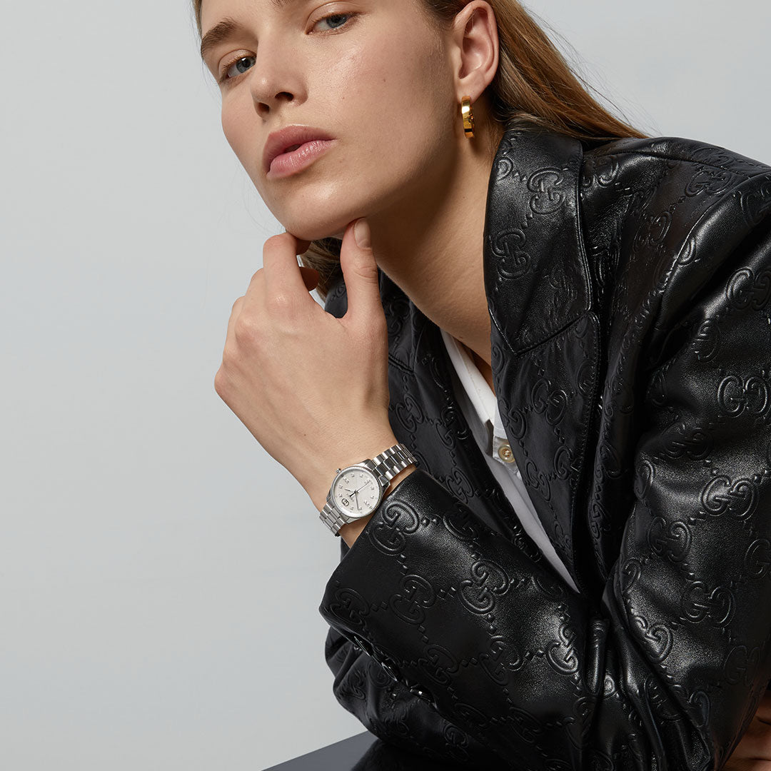 A woman in a black embossed leather blazer and gold hoop earrings poses with her hand near her chin, wearing a GUCCI Watch—Gucci G-Timeless Quartz 29mm—against a neutral background.