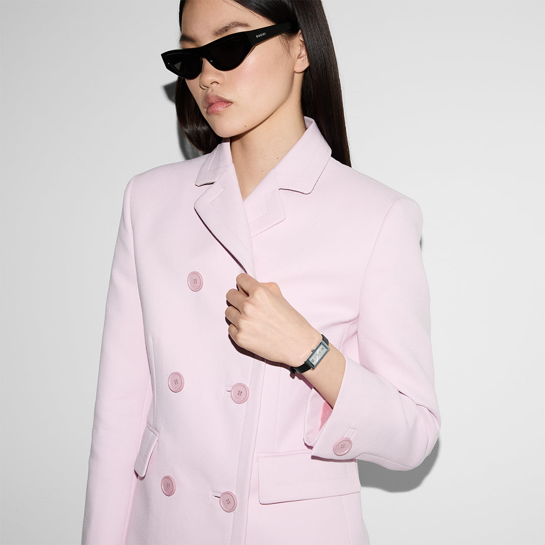 A person wearing cat-eye sunglasses and a light pink double-breasted coat holds the lapel with one hand. They have long, dark hair and are wearing a GUCCI Watch from the G-Frame collection, specifically the Gucci G-Frame Quartz 14 X 34mm Watch with a pink dial. The background is plain white.