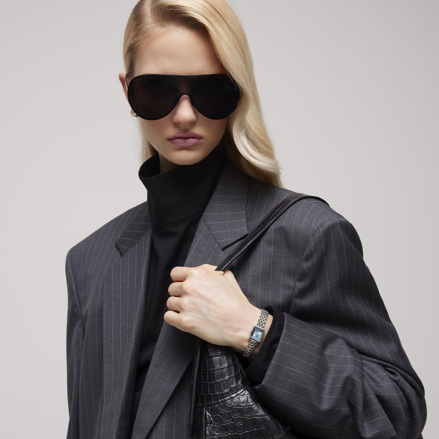 A woman in a pinstripe blazer, black turtleneck, and large dark sunglasses wears a GUCCI Watch G-Frame Quartz 14mm x 18mm with a stainless steel bracelet and holds a black textured handbag, exuding style and confidence.
