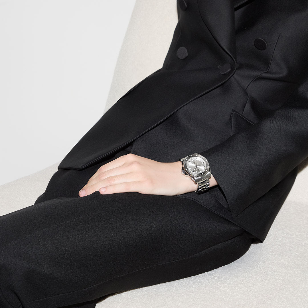 A person in a black suit sits on a light-colored chair, one hand on their lap, wearing a GUCCI Watch—Gucci Dive Automatic 40mm.