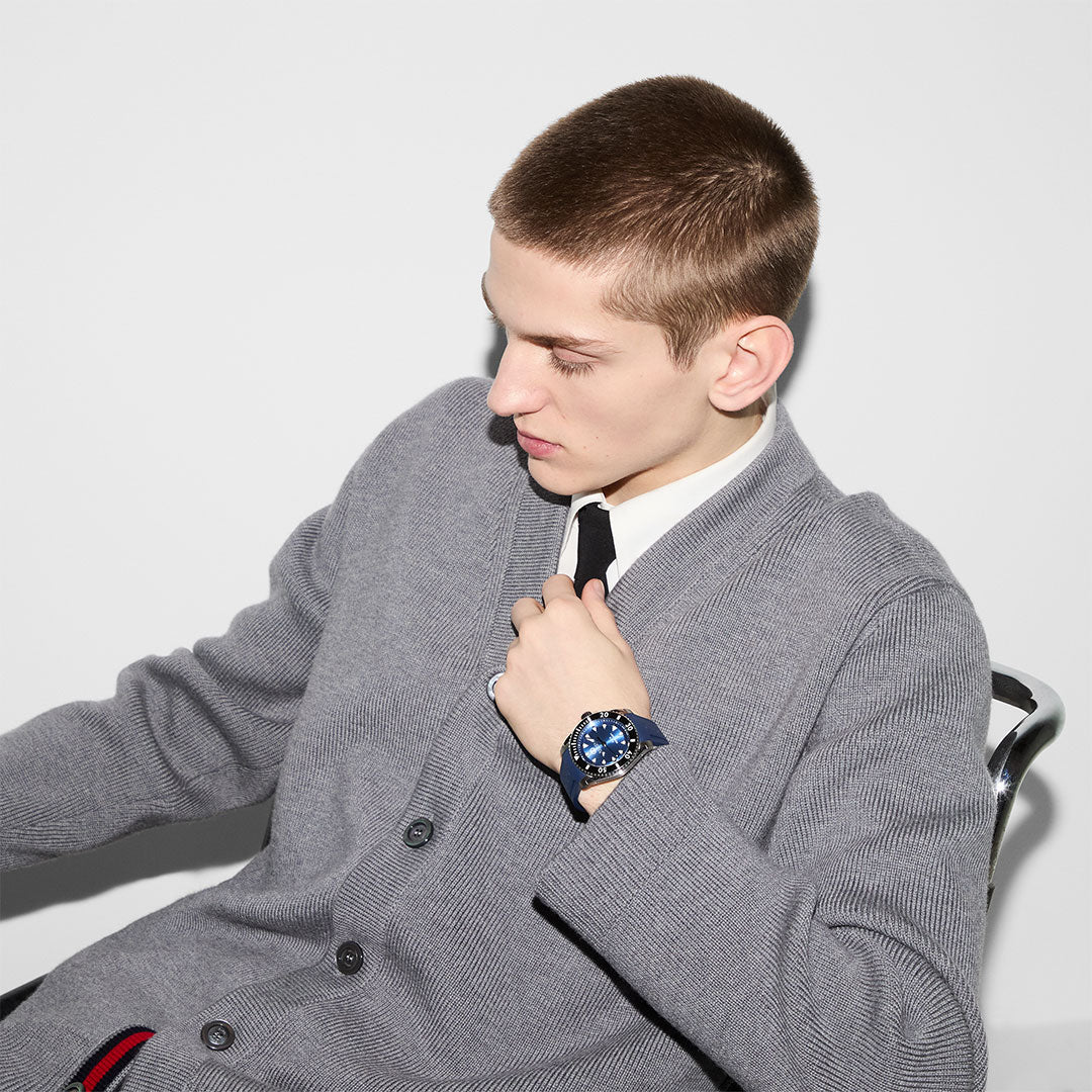 A person with short hair sits on a chair, wearing a gray cardigan over a white shirt and black tie. They hold their collar with one hand, revealing a stylish GUCCI Watch's Gucci Dive Automatic 40mm Watch with a blue dial featuring the Interlocking G, while looking down to their left against a light gray background.