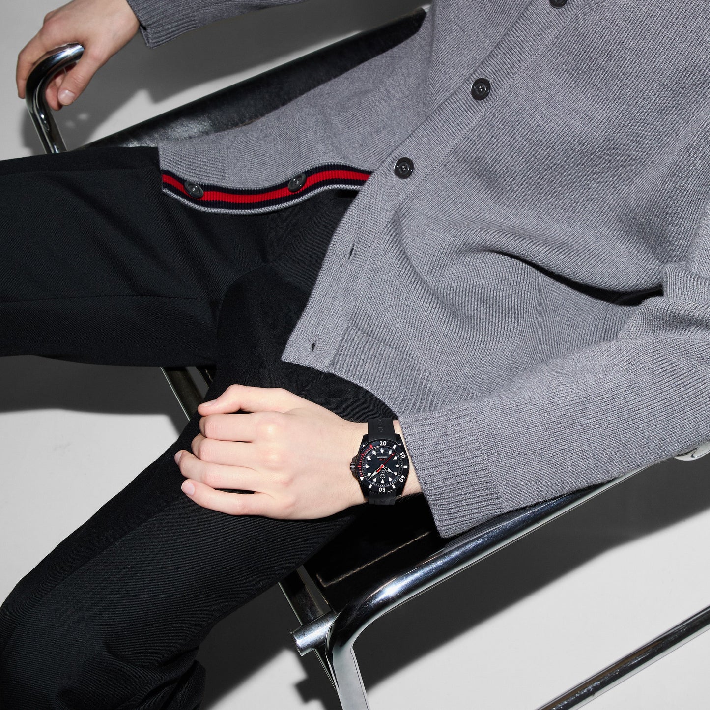 A person sits on a metallic, minimalist chair, wearing a gray cardigan accented with black and red stripes, paired with black pants. On their wrist is the elegant GUCCI Watch's Gucci Dive Black Automatic 40mm Watch, boasting a sleek black dial encased in stainless steel, enhancing their ensemble.